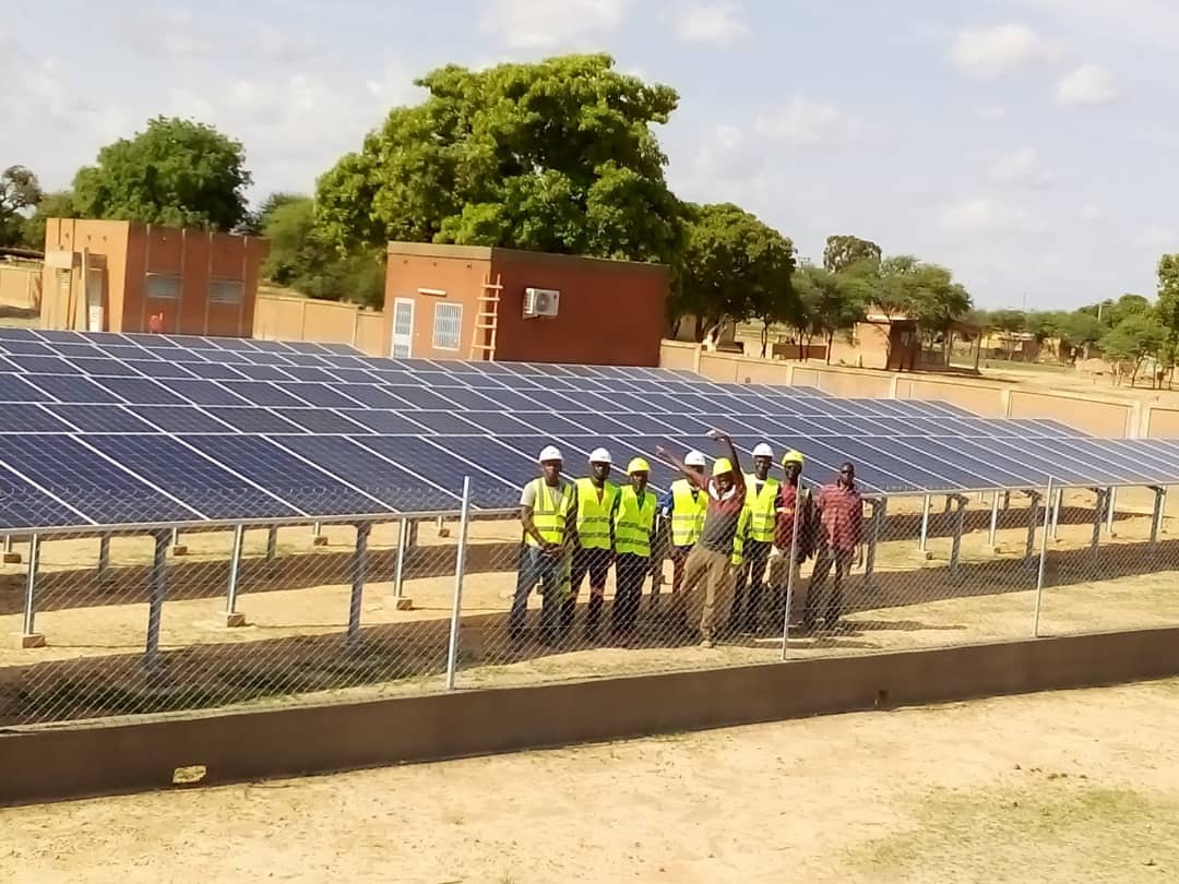 Installation crew in hard hats and hi-vis vests standing beside a ground-mounted solar array at a walled compound.