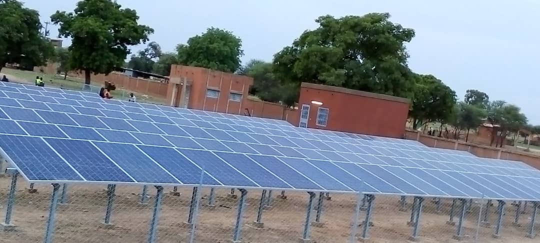Long row of ground-mounted solar panels on steel frames in a fenced compound.