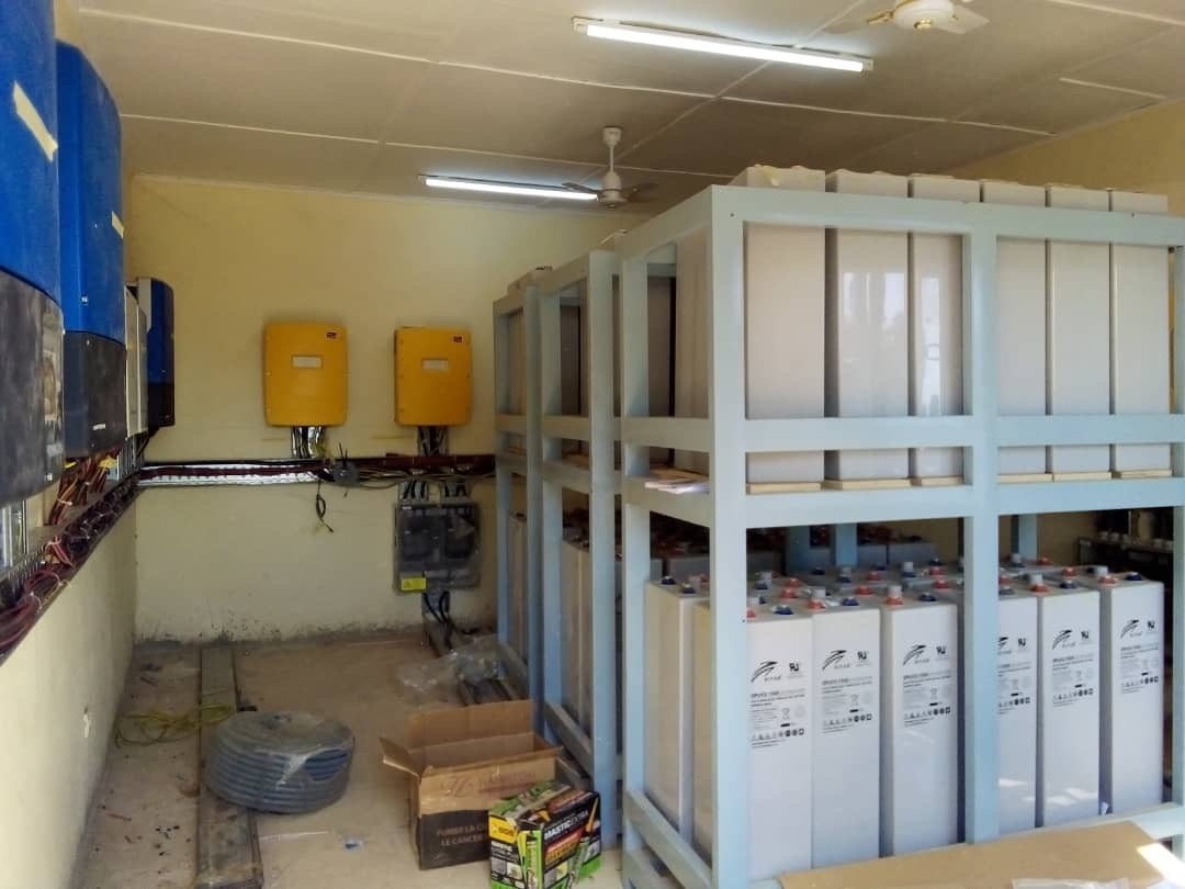 Wall-mounted inverters and charge controllers wired to a racked bank of lead-acid batteries in an equipment room.
