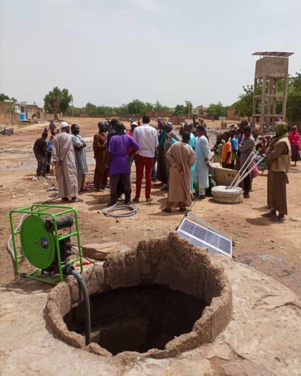 Solar surface pump set at the mouth of a lined open well, with a crowd of villagers and a water tower behind.