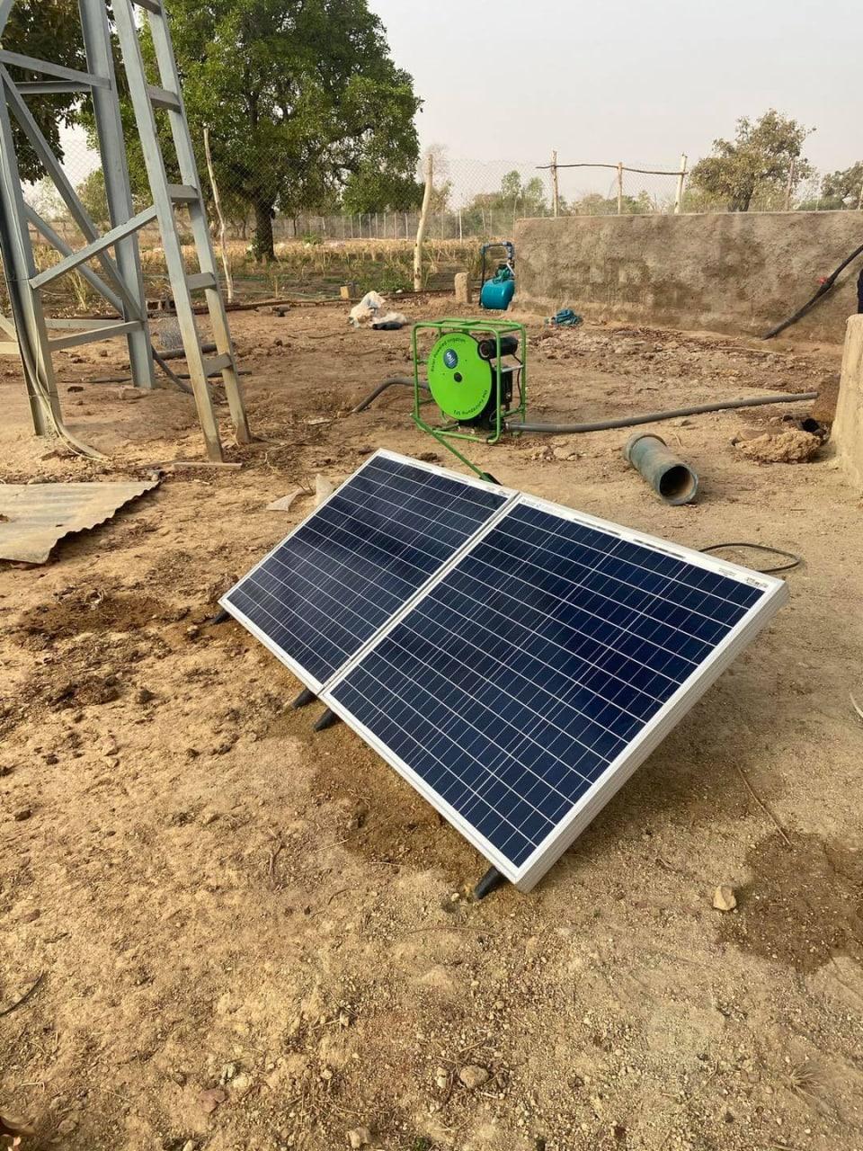 Solar panel pair propped on dry ground next to a green-framed pump at a walled water point.