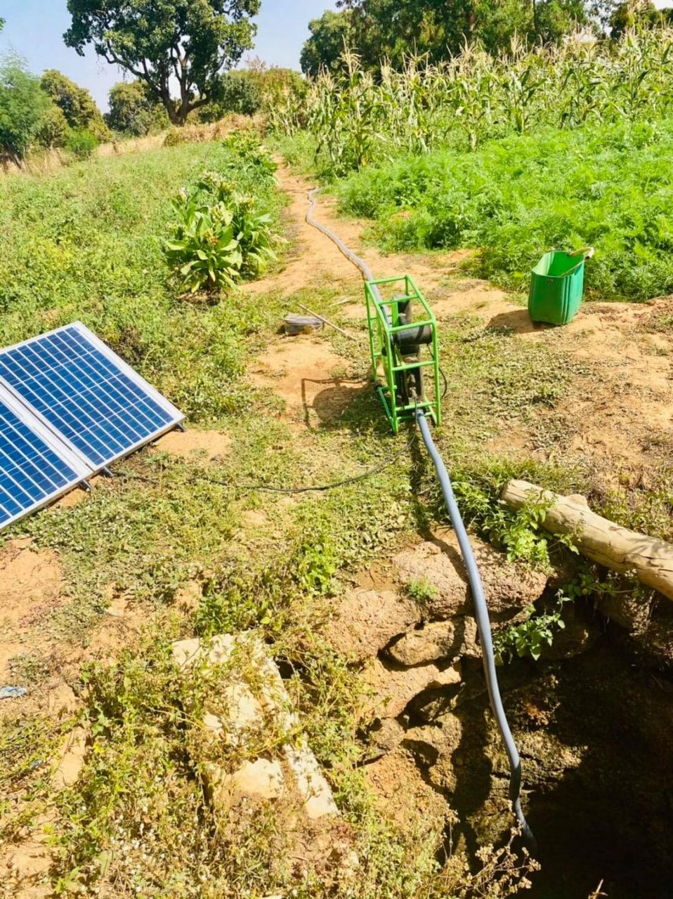 A solar surface pump in a green frame beside two folding solar panels, drawing water through a hose from a low stream bed up to a green market garden.