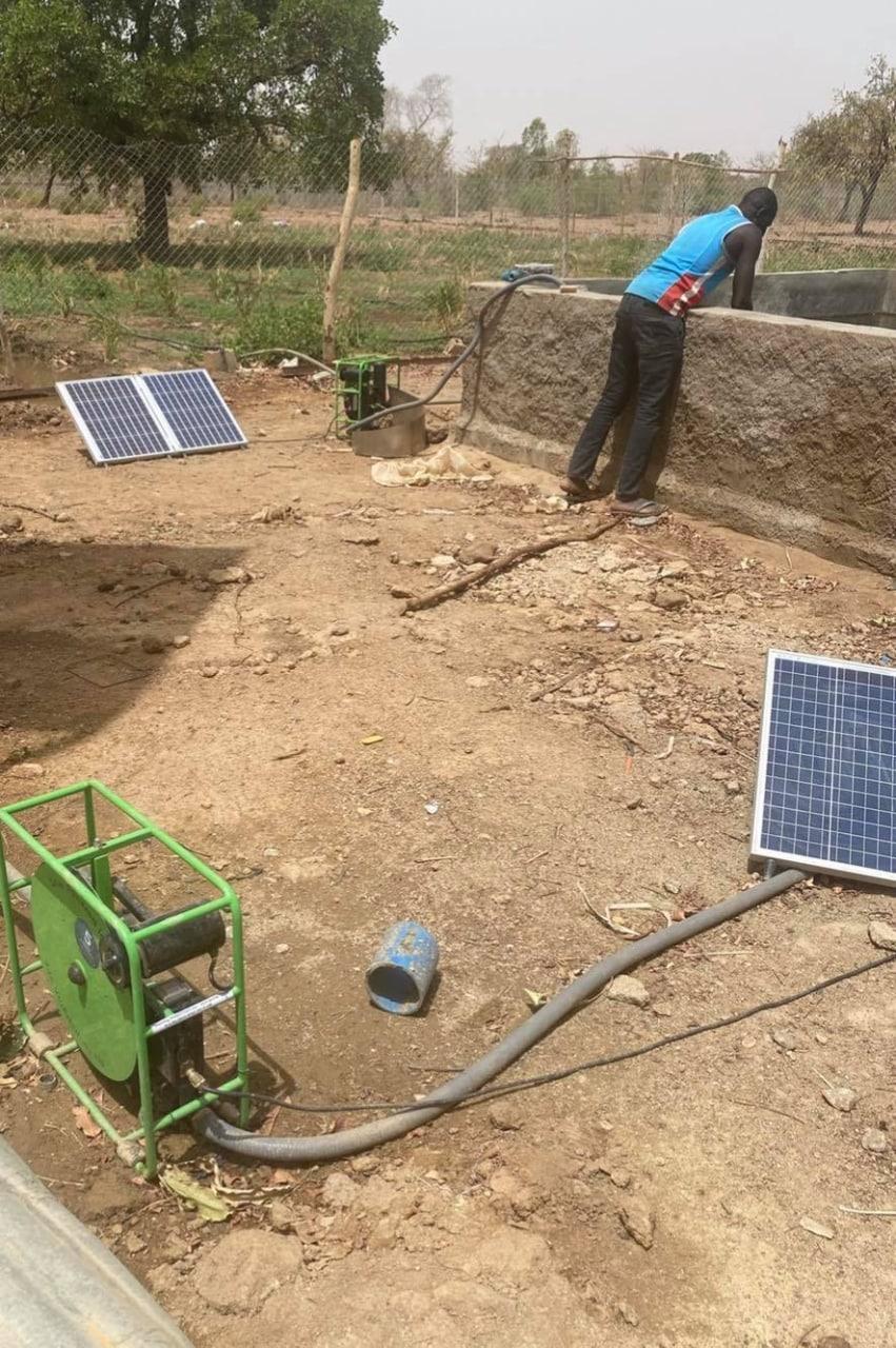 A man leaning over the wall of a concrete reservoir guiding a discharge hose into it, fed by a green-framed solar pump and two panels set on bare ground nearby.
