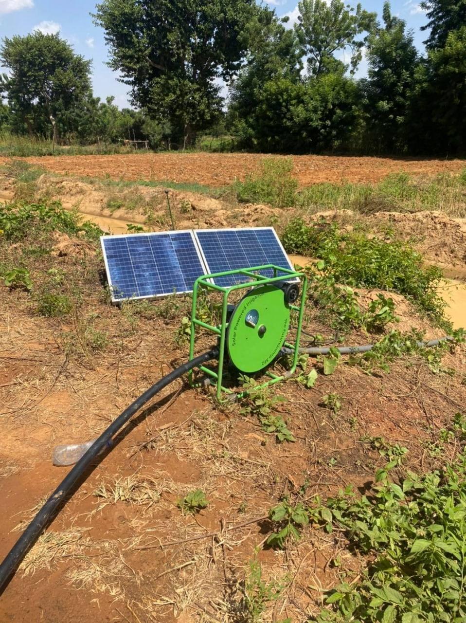 Portable solar surface pump in a green frame beside a folding solar panel at the edge of a field.