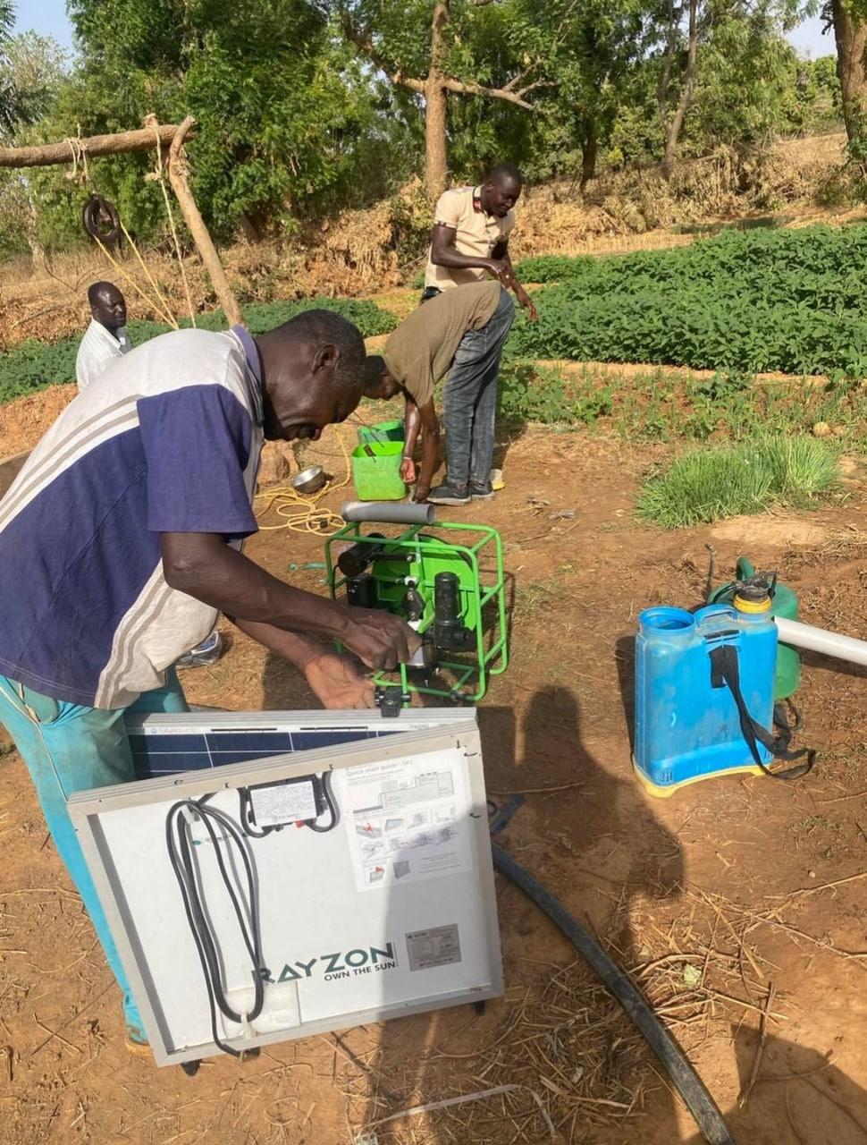 Three men setting up a solar surface pump and panel on bare ground at the edge of a market garden.